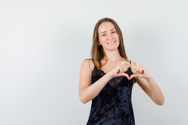 Young female showing heart gesture in black singlet and looking joyful.