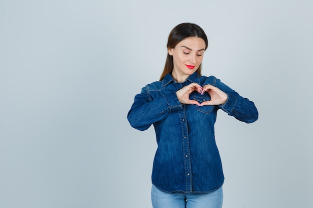 Foto gratuita giovane femmina che mostra il gesto del cuore in camicia di jeans e jeans e sembra carina