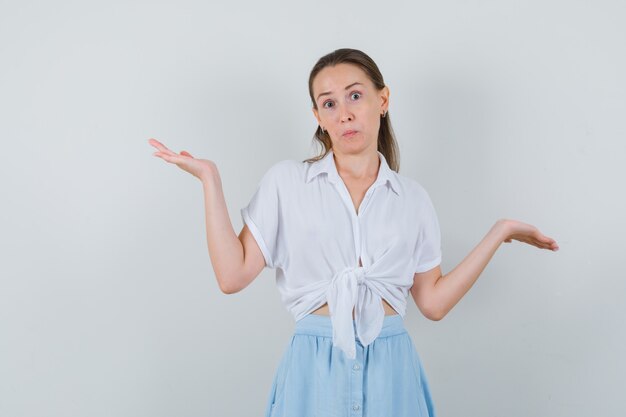 Young female showing helpless gesture in blouse and skirt and looking confused