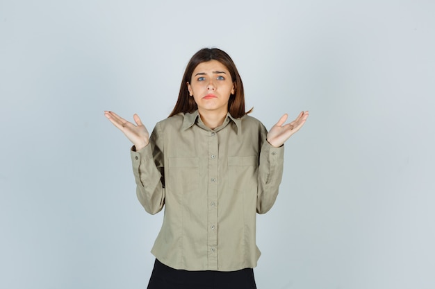 Young female showing helpless gesture, curving lips in shirt, skirt and looking puzzled , front view.