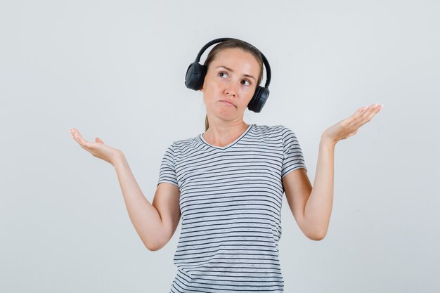 Young female showing helpless gesture in striped t-shirt, headphones , front view.