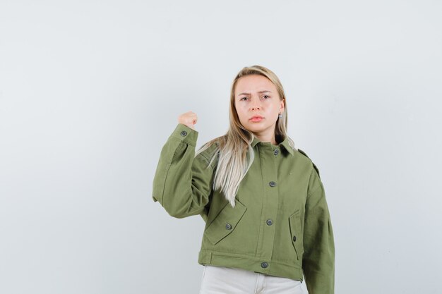 緑のジャケット、ジーンズで彼女の腕の力を示し、真剣に見える若い女性、正面図。