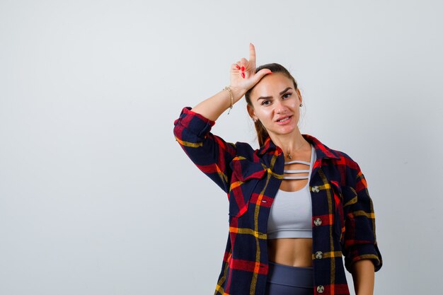 Young female showing loser gesture in crop top, checkered shirt and looking serious. front view.