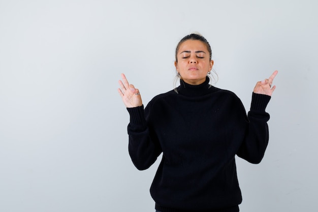 Young female showing meditation gesture in turtleneck sweater and looking peaceful. front view.
