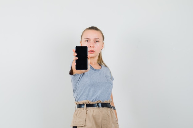 Молодая женщина показывает мобильный телефон в футболке, штанах и выглядит серьезным, вид спереди.
