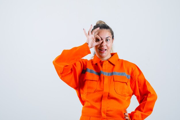 Young female showing ok gesture over eye in worker uniform and looking joyful. front view.