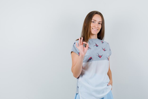 Tシャツ、スカートで大丈夫なジェスチャーを示し、陽気に見える若い女性、正面図。