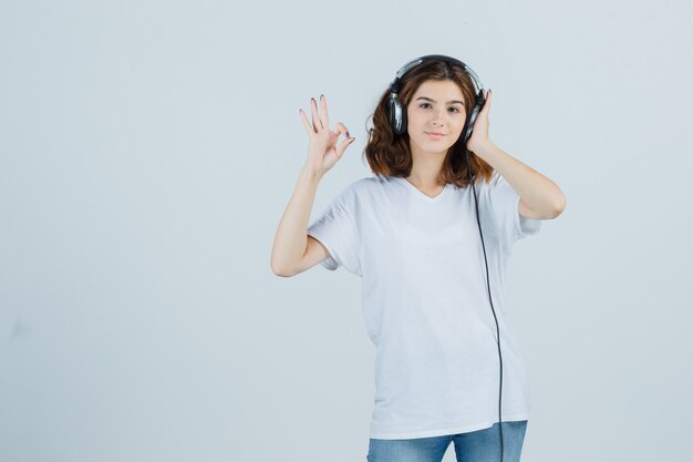 흰색 t- 셔츠에 헤드폰으로 음악을 듣고 유쾌한 찾고있는 동안 확인 제스처를 보여주는 젊은 여성. 전면보기.