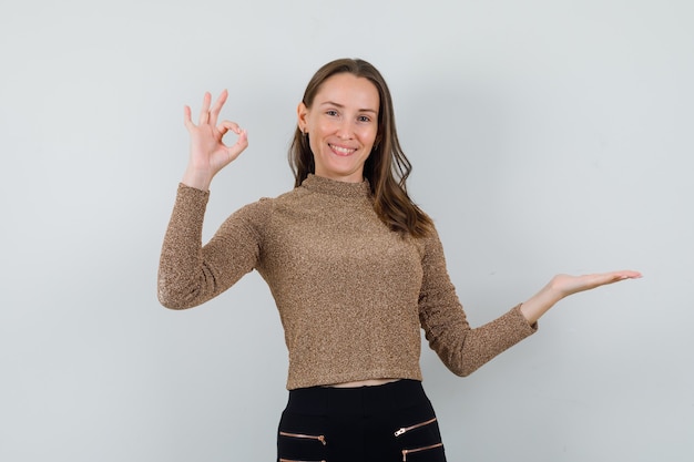 Young female showing ok gesture while raising hand up in golden blouse and looking glad. front view.