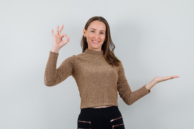 Young female showing ok gesture while raising hand up in golden blouse and looking glad. front view.