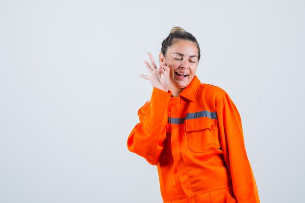 Young female showing ok gesture while winking in worker uniform and looking happy , front view.