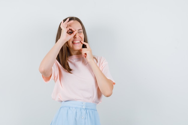 若い女性は、Tシャツ、スカート、おかしな顔でOKサインを示しています。正面図。