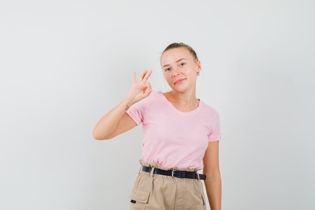 Giovane femmina che mostra segno giusto in t-shirt, pantaloni e sembra allegra, vista frontale.