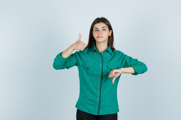 Young female showing opposite thumbs, curving lips in green shirt and looking indecisive. front view.
