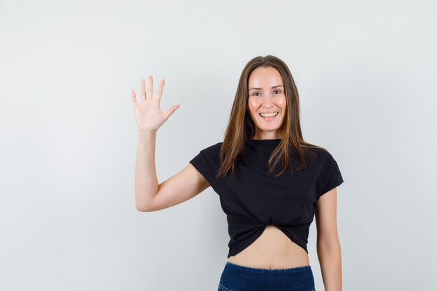 Young female showing palm in black blouse, pants and looking cheerful