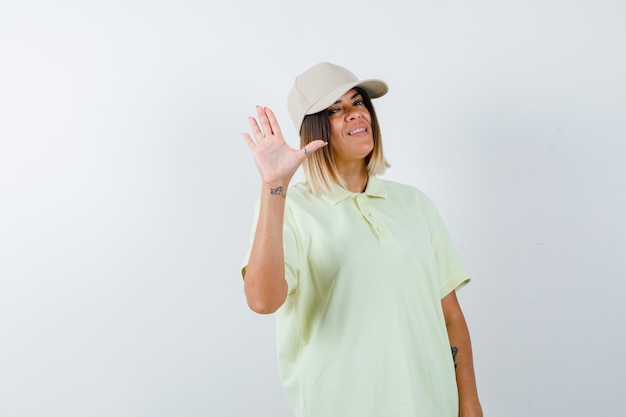 T- 셔츠, 모자에 인사 하 고 즐거운 찾고 손바닥을 보여주는 젊은 여성. 전면보기.