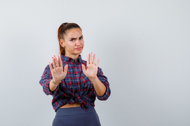 Young female showing palms in surrender gesture in checkered shirt, pants and looking serious , front view.