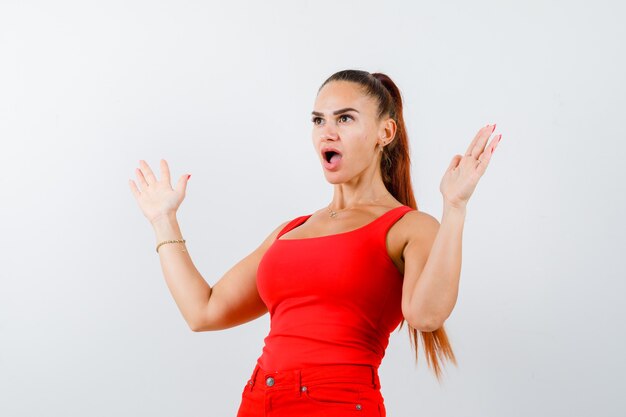 Young female showing palms in surrender gesture in red tank top, pants and looking puzzled , front view.