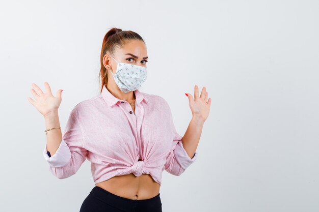 Young female showing palms in surrender gesture in shirt, pants, medical mask and looking helpless. front view.