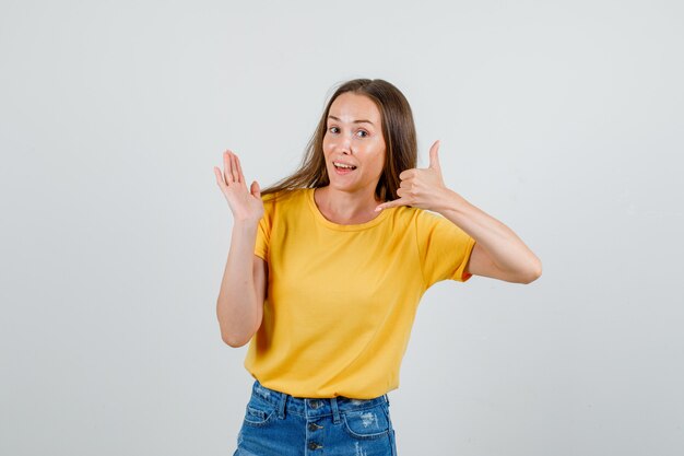 Tシャツ、ショートパンツ、陽気に見える開いた手のひらで電話のサインを示す若い女性