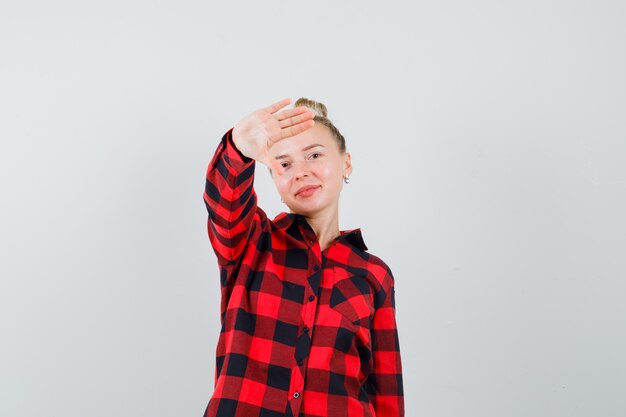 Young female showing refusal gesture politely in checked shirt and looking glad. front view.