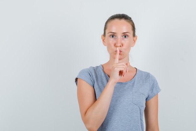 Young female showing silence gesture in grey t-shirt and looking careful. front view.