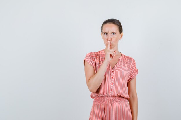 Young female showing silence gesture in striped dress and looking serious. front view.