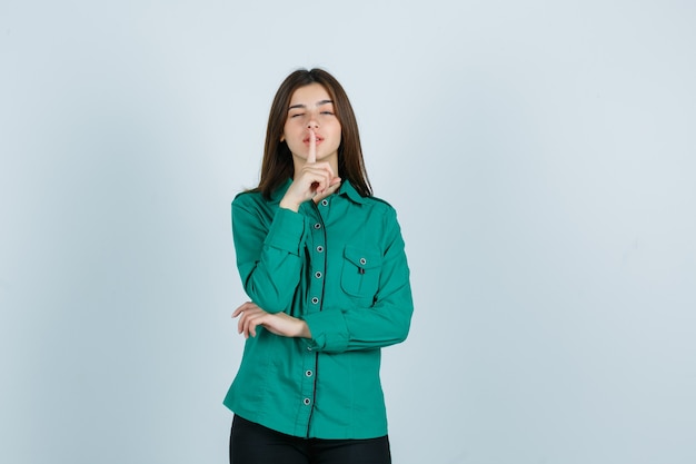 Young female showing silence gesture while blinking in green shirt and looking confident , front view.