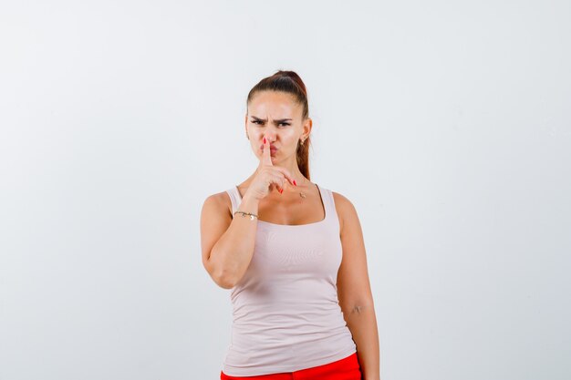 Young female showing silence gesture in white tank top, pants and looking serious. front view.