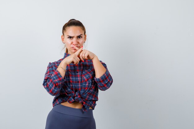 Молодая женщина показывает жест молчания со скрещенными пальцами в клетчатой рубашке, штанах и выглядит серьезно. передний план.