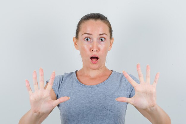 Young female showing stop gesture in grey t-shirt and looking scared , front view.