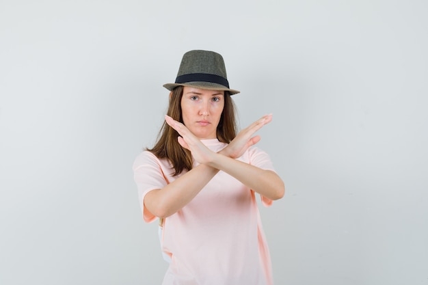 Молодая женщина показывает жест остановки в розовой футболке, шляпе и выглядит серьезным, вид спереди.