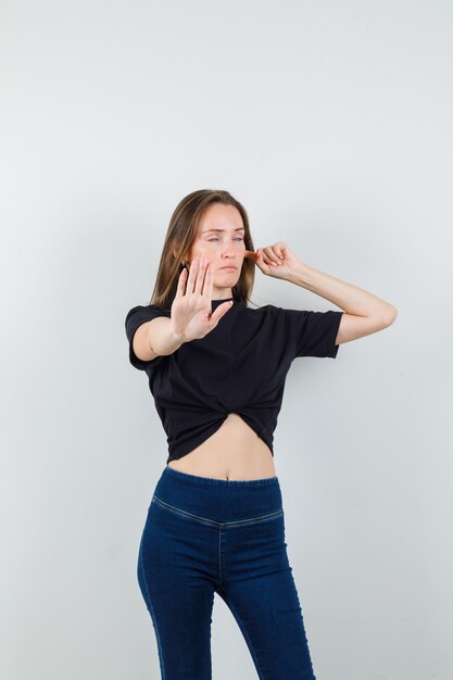 Молодая женщина показывает жест стоп, затыкая ухо в черной блузке