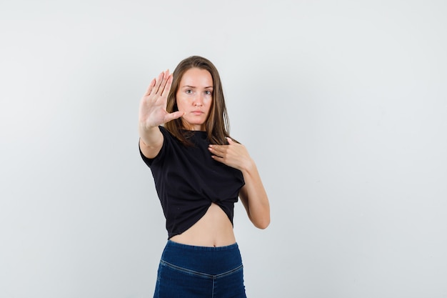 Young female showing stop gesture while pointing at herself in black blouse, pants and looking serious.