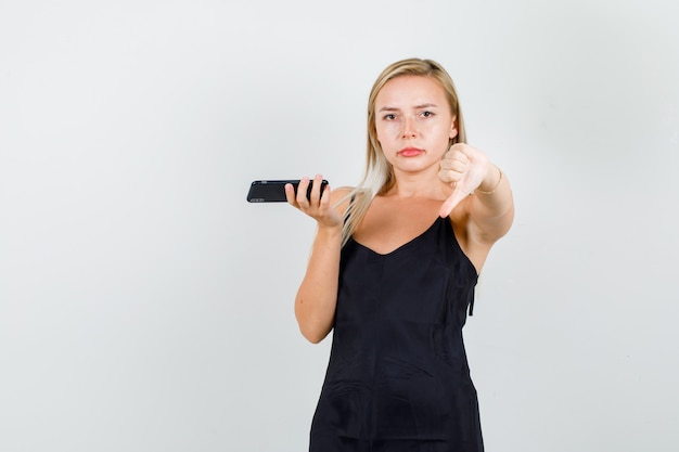 검은 색 단일 선에 스마트 폰을 들고 불만족스럽게 보는 동안 엄지 손가락을 아래로 보여주는 젊은 여성