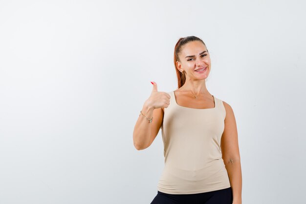 Young female showing thumb up in beige tank top and looking glad , front view.