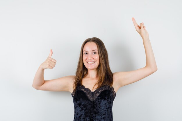 Young female showing thumb up, gesturing with two fingers in black singlet and looking cheerful.