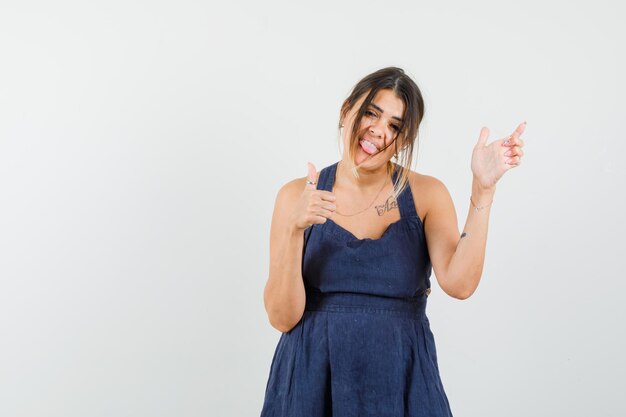 Young female showing thumb up, pointing away, sticking out tongue in dark blue dress