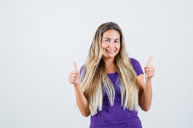 Молодая женщина показывает палец вверх в фиолетовой футболке и выглядит оптимистично, вид спереди.
