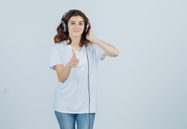 흰색 티셔츠, 청바지에 헤드폰으로 음악을 즐기고 행복을 찾는 동안 엄지 손가락을 보여주는 젊은 여성. 전면보기.