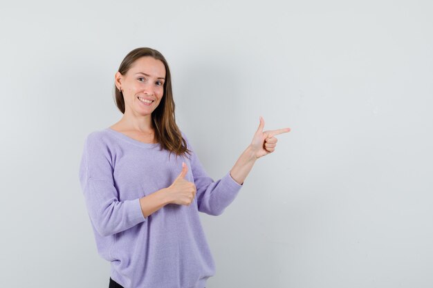 Молодая женщина показывает палец вверх, указывая в сторону в сиреневой блузке и выглядит позитивно. передний план.