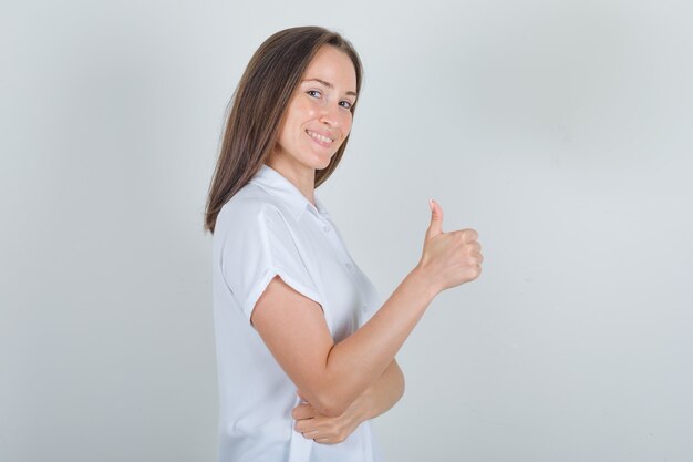 Young female showing thumb up in white shirt and looking pleased.