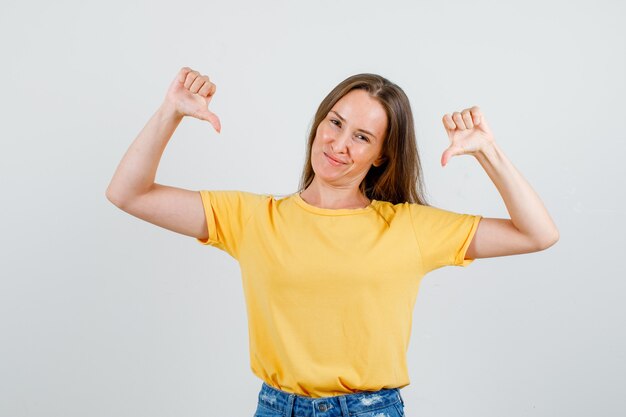 Young female showing thumbs down in t-shirt, shorts and looking displeased. front view.