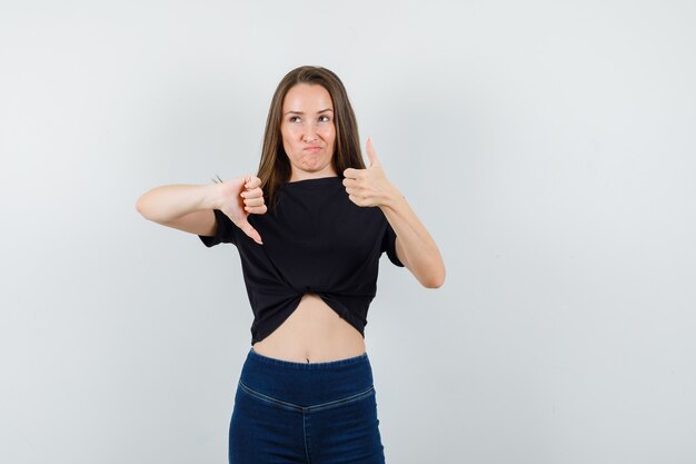 Young female showing thumbs up and down in black blouse, pants and looking hesitant
