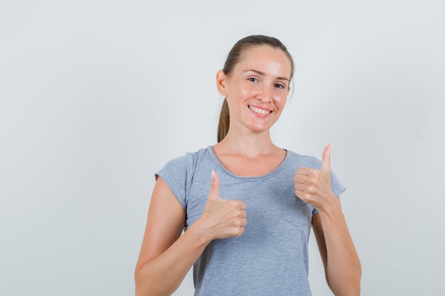 Young female showing thumbs up in grey t-shirt and looking happy. front view.