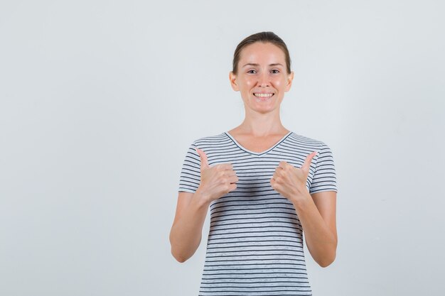 Young female showing thumbs up in striped t-shirt and looking cheerful. front view.