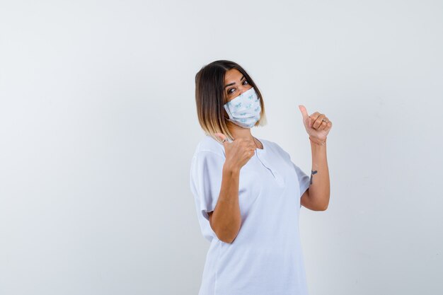 Young female showing thumbs up in t-shirt, mask and looking confident , front view.