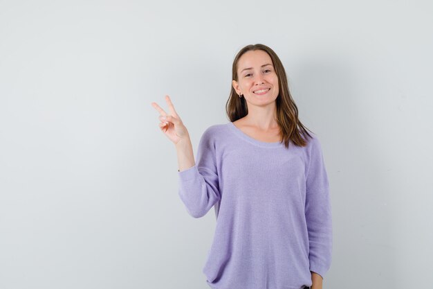 Молодая женщина показывает знак V в сиреневой блузке и выглядит радостной