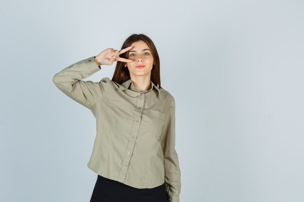 Young female showing V-sign near eye in shirt, skirt and looking confident , front view.