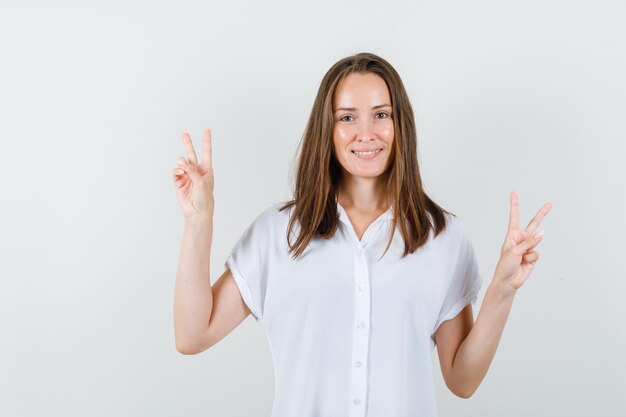 Young female showing victory gesture while smiling in white blouse and looking pleased